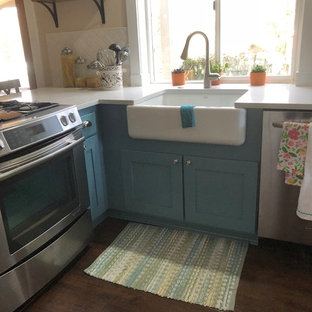 Aménagement d'une arrière-cuisine bord de mer en L de taille moyenne avec un évier de ferme, un placard à porte shaker, des portes de placard turquoises, un plan de travail en quartz, une crédence blanche, une crédence en mosaïque, un électroménager en acier inoxydable, sol en stratifié, une péninsule, un sol marron et un plan de travail blanc.