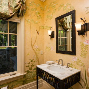 Imagen de aseo tradicional, grande, con lavabo bajoencimera, armarios tipo mueble, puertas de armario negras, paredes amarillas, suelo de travertino, encimera de mármol, suelo beige y encimeras blancas