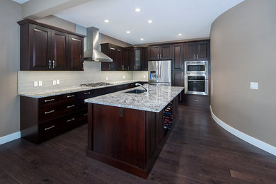 Large transitional l-shaped eat-in kitchen in Calgary with an undermount sink, dark wood cabinets, granite benchtops, white splashback, glass tile splashback, stainless steel appliances, dark hardwood floors, with island and raised-panel cabinets.
