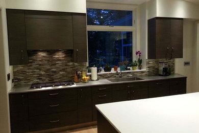 Example of a trendy light wood floor and brown floor kitchen design in Other with an undermount sink, flat-panel cabinets, dark wood cabinets, stainless steel countertops, multicolored backsplash, matchstick tile backsplash, paneled appliances and an island