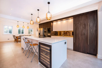 This is an example of a large contemporary grey and brown galley open plan kitchen in London with a submerged sink, flat-panel cabinets, dark wood cabinets, quartz worktops, metallic splashback, mirror splashback, stainless steel appliances, ceramic flooring, an island, multi-coloured floors, white worktops, a vaulted ceiling and feature lighting.