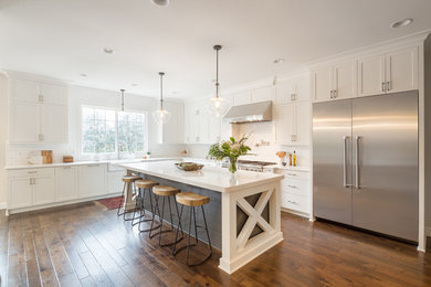 This is an example of a transitional l-shaped kitchen in Portland with a farmhouse sink, shaker cabinets, white cabinets, white splashback, subway tile splashback, stainless steel appliances, with island and dark hardwood floors.
