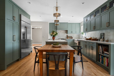 Transitional u-shaped light wood floor and brown floor kitchen photo in Detroit with a farmhouse sink, shaker cabinets, green cabinets, quartz countertops, white backsplash, ceramic backsplash, stainless steel appliances, an island and white countertops