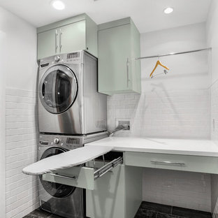 Diseño de cuarto de lavado lineal, retro, de tamaño medio, con armarios con paneles lisos, encimera de acrílico, paredes blancas, suelo de baldosas de porcelana, lavadora y secadora apiladas, suelo gris y puertas de armario verdes