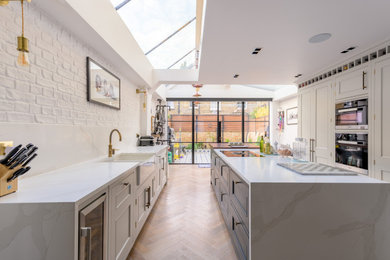 Photo of a large traditional galley open plan kitchen in London with a built-in sink, recessed-panel cabinets, beige cabinets, marble worktops, white splashback, marble splashback, black appliances, light hardwood flooring, an island, brown floors, white worktops, a vaulted ceiling and feature lighting.