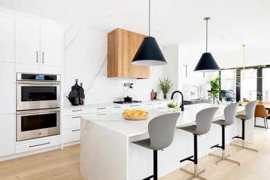 This is an example of a large modern l-shaped eat-in kitchen in Other with flat-panel cabinets, white cabinets, solid surface benchtops, white splashback, engineered quartz splashback, stainless steel appliances, light hardwood floors, with island, brown floor and white benchtop.