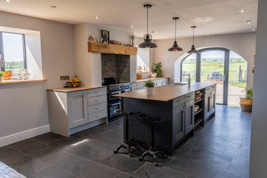 Photo of a traditional galley kitchen/diner in Other with a belfast sink, shaker cabinets, grey cabinets, laminate countertops, black appliances, an island, black floors and brown worktops.