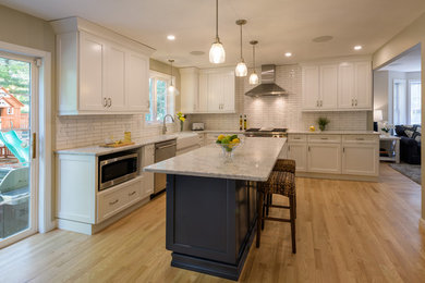 Mid-sized transitional l-shaped eat-in kitchen in Boston with white splashback, subway tile splashback, with island, white benchtop, a farmhouse sink, raised-panel cabinets, white cabinets, quartzite benchtops, stainless steel appliances and medium hardwood floors.