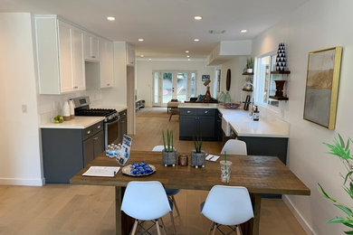 Design ideas for a mid-sized transitional galley open plan kitchen in Los Angeles with a farmhouse sink, shaker cabinets, blue cabinets, solid surface benchtops, stainless steel appliances, light hardwood floors, a peninsula, beige floor and white benchtop.
