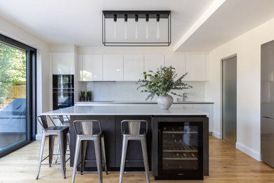 Photo of a modern galley kitchen in Sussex with a submerged sink, flat-panel cabinets, white cabinets, white splashback, black appliances, medium hardwood flooring, an island, brown floors and white worktops.