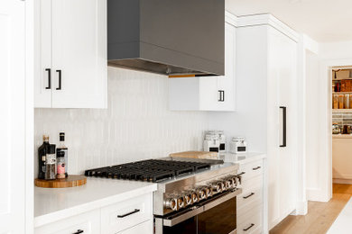 Inspiration for a large transitional single-wall medium tone wood floor and brown floor open concept kitchen remodel in Toronto with a farmhouse sink, shaker cabinets, white cabinets, quartz countertops, white backsplash, ceramic backsplash, stainless steel appliances, an island and white countertops