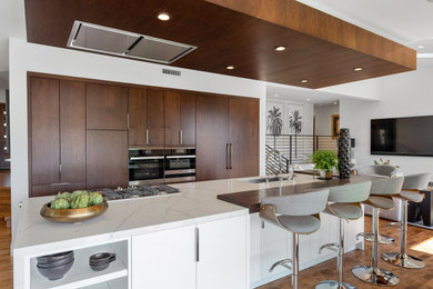 This is an example of an expansive contemporary open plan kitchen in Los Angeles with a built-in sink, flat-panel cabinets, dark wood cabinets, engineered stone countertops, medium hardwood flooring, an island, brown floors and white worktops.