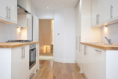 Inspiration for a small contemporary galley enclosed kitchen in London with a built-in sink, white cabinets, wood worktops, white splashback, stainless steel appliances, light hardwood flooring, no island, beige floors and brown worktops.