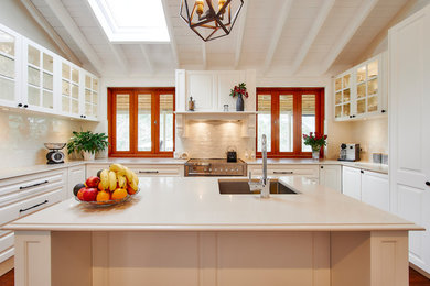 This is an example of a large traditional u-shaped kitchen in Sydney with an undermount sink, recessed-panel cabinets, white cabinets, quartz benchtops, white splashback, travertine splashback, white appliances, medium hardwood floors, with island and multi-coloured floor.