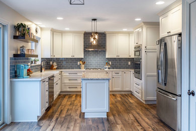 Example of a mid-sized trendy u-shaped medium tone wood floor and brown floor eat-in kitchen design in Other with a farmhouse sink, shaker cabinets, white cabinets, quartz countertops, blue backsplash, glass tile backsplash, stainless steel appliances, an island and white countertops