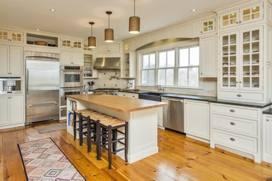 Inspiration for a mid-sized transitional l-shaped medium tone wood floor and brown floor eat-in kitchen remodel in Portland Maine with a farmhouse sink, recessed-panel cabinets, white cabinets, soapstone countertops, white backsplash, stone tile backsplash, stainless steel appliances, an island and black countertops