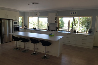 Large modern galley open plan kitchen in Sunshine Coast with a double-bowl sink, white cabinets, laminate benchtops, grey splashback, ceramic splashback, stainless steel appliances, laminate floors, with island, beige floor and white benchtop.