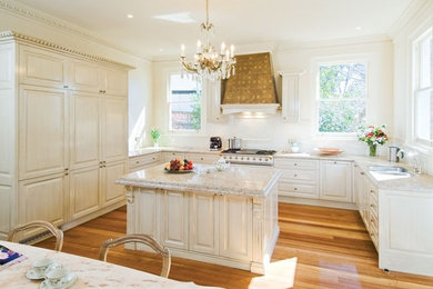 Photo of a large traditional u-shaped open plan kitchen in Melbourne with a double-bowl sink, raised-panel cabinets, white cabinets, marble benchtops, white splashback, ceramic splashback, white appliances, medium hardwood floors and with island.