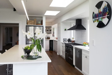 Inspiration for a traditional galley open plan kitchen in Wellington with an undermount sink, shaker cabinets, white cabinets, solid surface benchtops, white splashback, subway tile splashback, black appliances, medium hardwood floors, with island, brown floor and white benchtop.