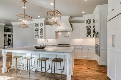 This is an example of a large transitional l-shaped eat-in kitchen in Oklahoma City with a farmhouse sink, flat-panel cabinets, white cabinets, marble benchtops, white splashback, subway tile splashback, stainless steel appliances, light hardwood floors, with island and brown floor.