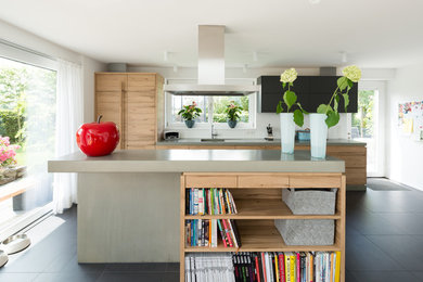 This is an example of a mid-sized contemporary single-wall open plan kitchen in Other with a single-bowl sink, flat-panel cabinets, light wood cabinets, concrete benchtops, white splashback, black appliances, ceramic floors, with island and grey floor.