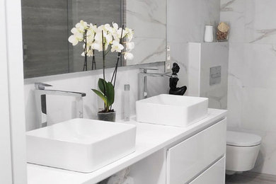 This is an example of a mid-sized transitional master bathroom in Bilbao with furniture-like cabinets, white cabinets, an open shower, a wall-mount toilet, black and white tile, ceramic tile, multi-coloured walls, travertine floors, solid surface benchtops, beige floor, a hinged shower door and white benchtops.