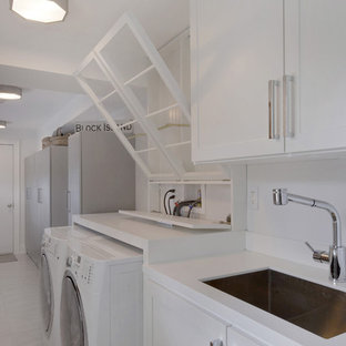 Diseño de cuarto de lavado lineal, minimalista, grande, con fregadero bajoencimera, armarios con paneles empotrados, puertas de armario blancas, encimera de acrílico, paredes blancas, suelo de baldosas de porcelana y lavadora y secadora juntas