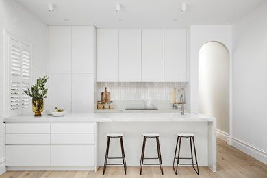 This is an example of a modern galley kitchen in Melbourne with an undermount sink, flat-panel cabinets, white cabinets, white splashback, panelled appliances, medium hardwood floors, a peninsula, brown floor and white benchtop.