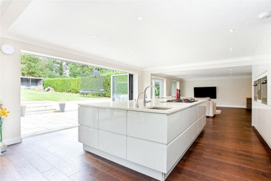 Large modern galley kitchen/diner in London with a double-bowl sink, flat-panel cabinets, beige cabinets, composite countertops, white splashback, an island and white worktops.