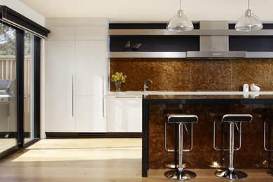 This is an example of a mid-sized beach style single-wall eat-in kitchen in Melbourne with flat-panel cabinets, white cabinets, brown splashback, granite benchtops, stone slab splashback, stainless steel appliances, light hardwood floors and with island.