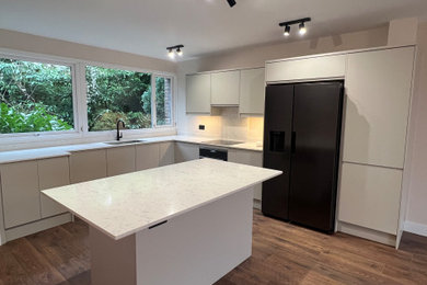 This is an example of a large kitchen in London with an integrated sink, flat-panel cabinets, grey cabinets, quartz worktops, white splashback, black appliances, an island and brown floors.