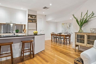 This is an example of a mid-sized contemporary galley open plan kitchen in Melbourne with a drop-in sink, flat-panel cabinets, white cabinets, quartz benchtops, metallic splashback, metal splashback, stainless steel appliances, light hardwood floors, with island, brown floor and black benchtop.