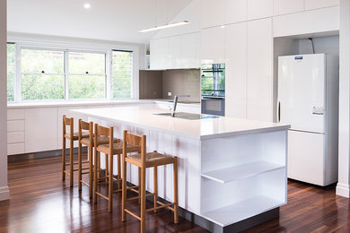Mid-sized modern galley open plan kitchen in Brisbane with a drop-in sink, flat-panel cabinets, white cabinets, marble benchtops, white splashback, glass sheet splashback, stainless steel appliances, medium hardwood floors, with island, brown floor and white benchtop.