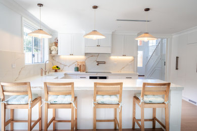 Mid-sized u-shaped kitchen pantry in Sydney with a farmhouse sink, recessed-panel cabinets, white cabinets, quartz benchtops, white splashback, engineered quartz splashback, white appliances, laminate floors, with island, brown floor and white benchtop.