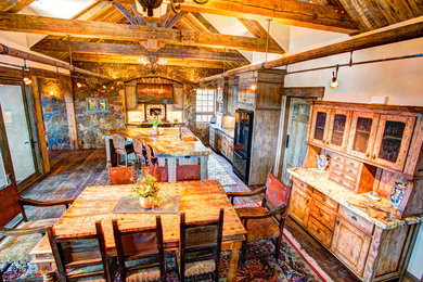 Large mountain style l-shaped dark wood floor eat-in kitchen photo in Other with a farmhouse sink, flat-panel cabinets, distressed cabinets, granite countertops, beige backsplash, stone tile backsplash, black appliances and an island
