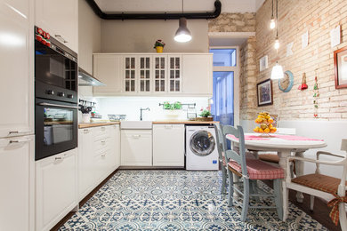Mid-sized transitional l-shaped eat-in kitchen in Alicante-Costa Blanca with a farmhouse sink, white cabinets, wood benchtops, white splashback, black appliances, ceramic floors, no island, glass-front cabinets and multi-coloured floor.