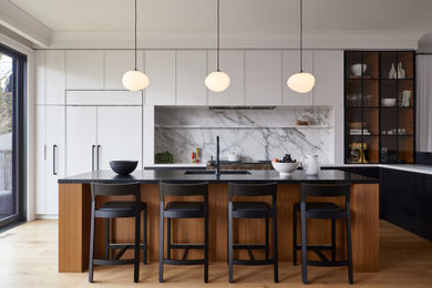 This is an example of a transitional l-shaped kitchen in Toronto with an undermount sink, flat-panel cabinets, white cabinets, white splashback, stone slab splashback, stainless steel appliances, medium hardwood floors, with island, brown floor and white benchtop.