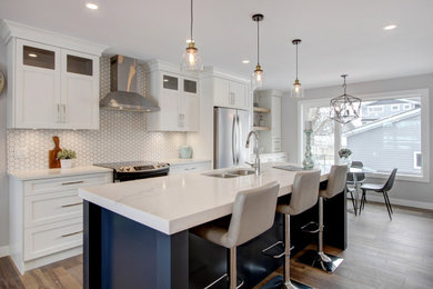 Inspiration for a mid-sized transitional galley open plan kitchen in Calgary with an undermount sink, shaker cabinets, white cabinets, quartz benchtops, white splashback, porcelain splashback, stainless steel appliances, medium hardwood floors, with island, brown floor and white benchtop.