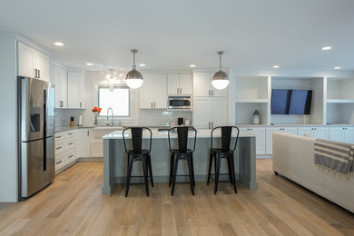 Inspiration for a mid-sized beach style l-shaped open plan kitchen in Grand Rapids with a farmhouse sink, shaker cabinets, white cabinets, quartz benchtops, white splashback, subway tile splashback, stainless steel appliances, light hardwood floors and with island.