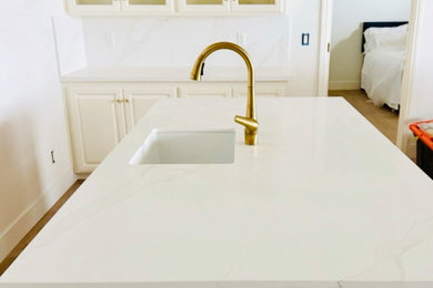 Example of a trendy medium tone wood floor kitchen design with an undermount sink, glass-front cabinets, white cabinets, quartzite countertops, quartz backsplash, stainless steel appliances and white countertops