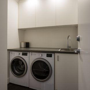 Photo of a small contemporary single-wall separated utility room in Auckland with a single-bowl sink, flat-panel cabinets, white cabinets, laminate countertops, white walls, laminate floors, a side by side washer and dryer and brown floors.