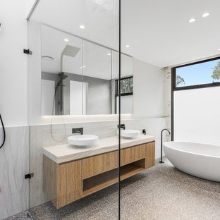 Cette image montre une douche en alcôve principale design de taille moyenne avec un placard à porte plane, des portes de placard en bois clair, une baignoire indépendante, un WC suspendu, un carrelage blanc, des carreaux de porcelaine, un mur blanc, sol en terrazzo, une vasque, un plan de toilette en marbre, un sol gris et un plan de toilette blanc.
