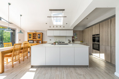 Photo of a large contemporary grey and white l-shaped open plan kitchen in Essex with a built-in sink, flat-panel cabinets, quartz worktops, beige splashback, glass sheet splashback, black appliances, porcelain flooring, an island, grey floors, white worktops and a vaulted ceiling.