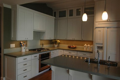 Example of a mid-sized trendy l-shaped medium tone wood floor open concept kitchen design in Other with a drop-in sink, recessed-panel cabinets, white cabinets, solid surface countertops, gray backsplash, stone slab backsplash, stainless steel appliances and an island