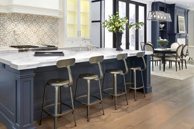 Mid-sized transitional l-shaped medium tone wood floor open concept kitchen photo in Chicago with a farmhouse sink, recessed-panel cabinets, white cabinets, marble countertops, white backsplash, mosaic tile backsplash, stainless steel appliances and an island