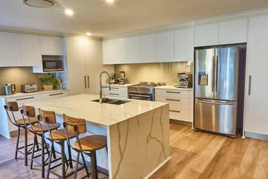 Photo of a mid-sized contemporary l-shaped open plan kitchen in Gold Coast - Tweed with a double-bowl sink, flat-panel cabinets, white cabinets, quartz benchtops, beige splashback, glass sheet splashback, stainless steel appliances, laminate floors, with island, brown floor and beige benchtop.