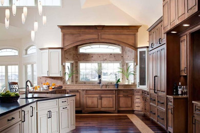 Example of a huge classic u-shaped dark wood floor, brown floor and vaulted ceiling open concept kitchen design in Other with an undermount sink, raised-panel cabinets, brown cabinets, quartz countertops, brown backsplash, travertine backsplash, paneled appliances, an island and black countertops