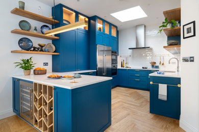Inspiration for a large modern u-shaped open plan kitchen in London with an integrated sink, shaker cabinets, blue cabinets, composite countertops, white splashback, terracotta splashback, stainless steel appliances, light hardwood flooring, an island, brown floors, white worktops and feature lighting.