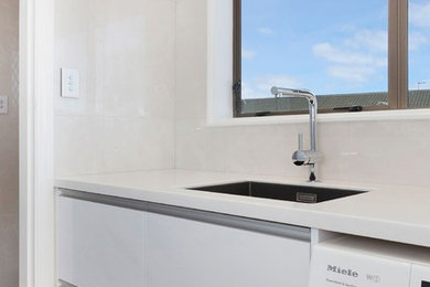 Photo of a mid-sized contemporary galley dedicated laundry room in Christchurch with an undermount sink, white cabinets, solid surface benchtops, grey walls, laminate floors, a side-by-side washer and dryer and black floor.