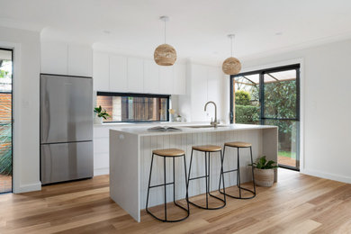 Mid-sized beach style galley open plan kitchen in Sydney with an undermount sink, flat-panel cabinets, white cabinets, quartz benchtops, window splashback, black appliances, medium hardwood floors, with island and white benchtop.